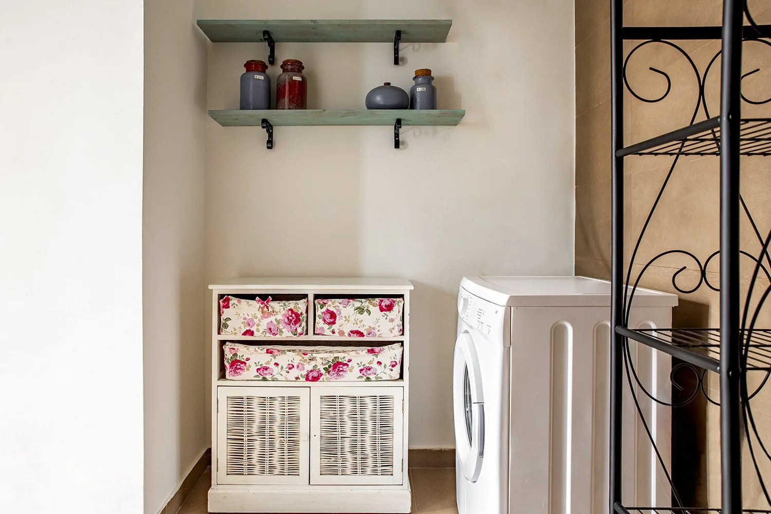 Zona de lavandería con una lavadora, un mueble floral y estantes en la pared.