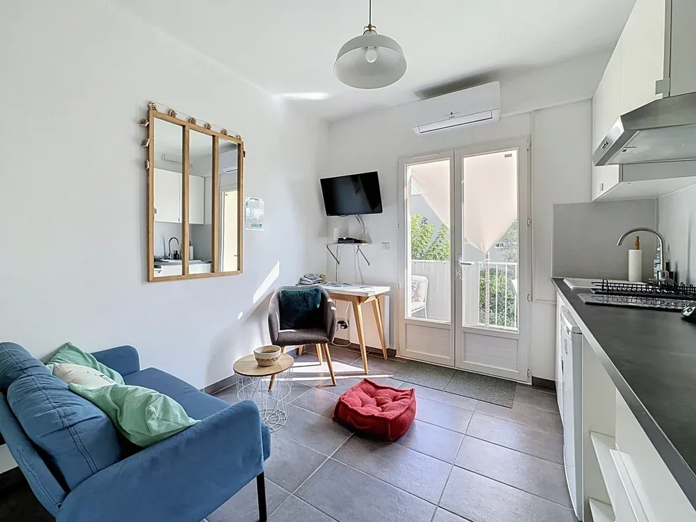 Una luminosa sala de estar se abre a una pequeña cocina, con un sofá azul y puertas de vidrio.