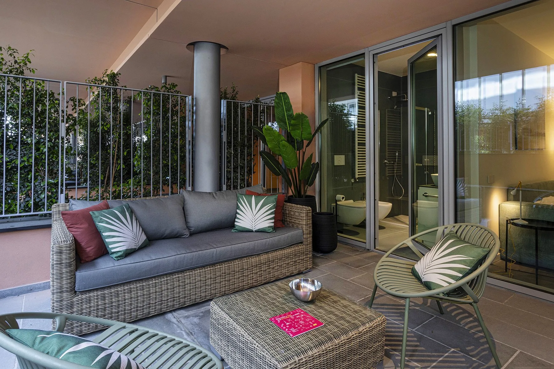 A furnished outdoor seating area beside glass doors, with potted plants and a leafy view.