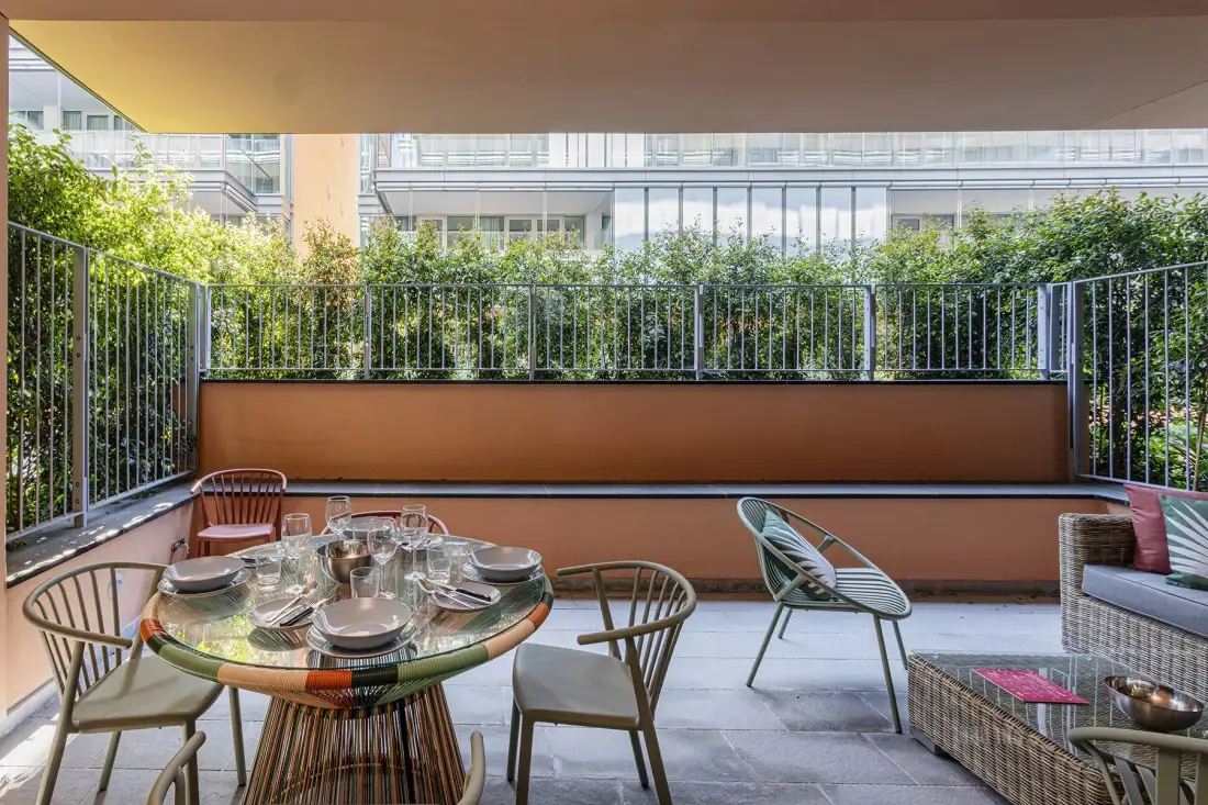 A covered outdoor patio has a round dining table, seating, and leafy plants.