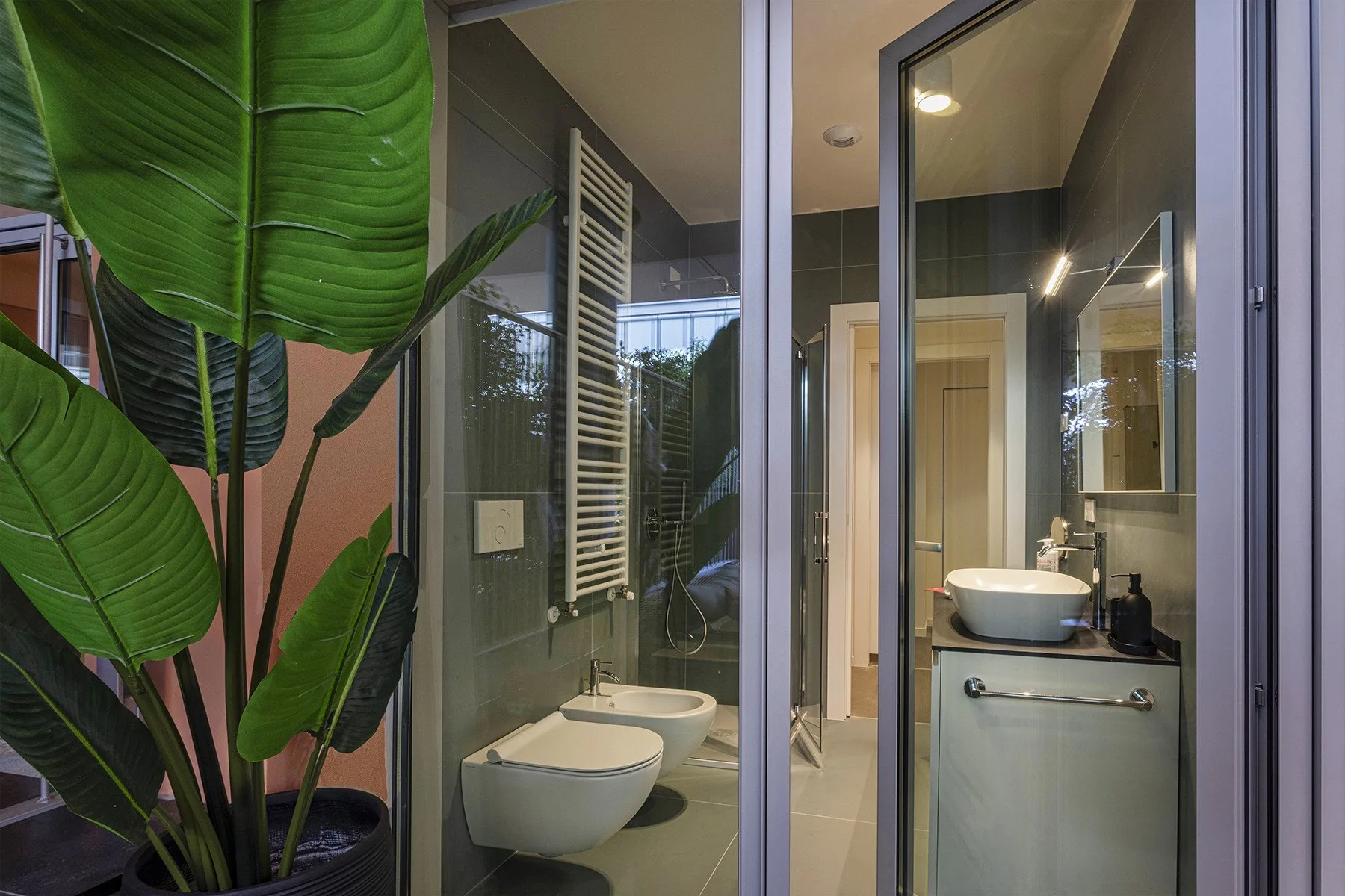 A modern bathroom with a glass shower, wall-mounted toilet, and large leafy plant.