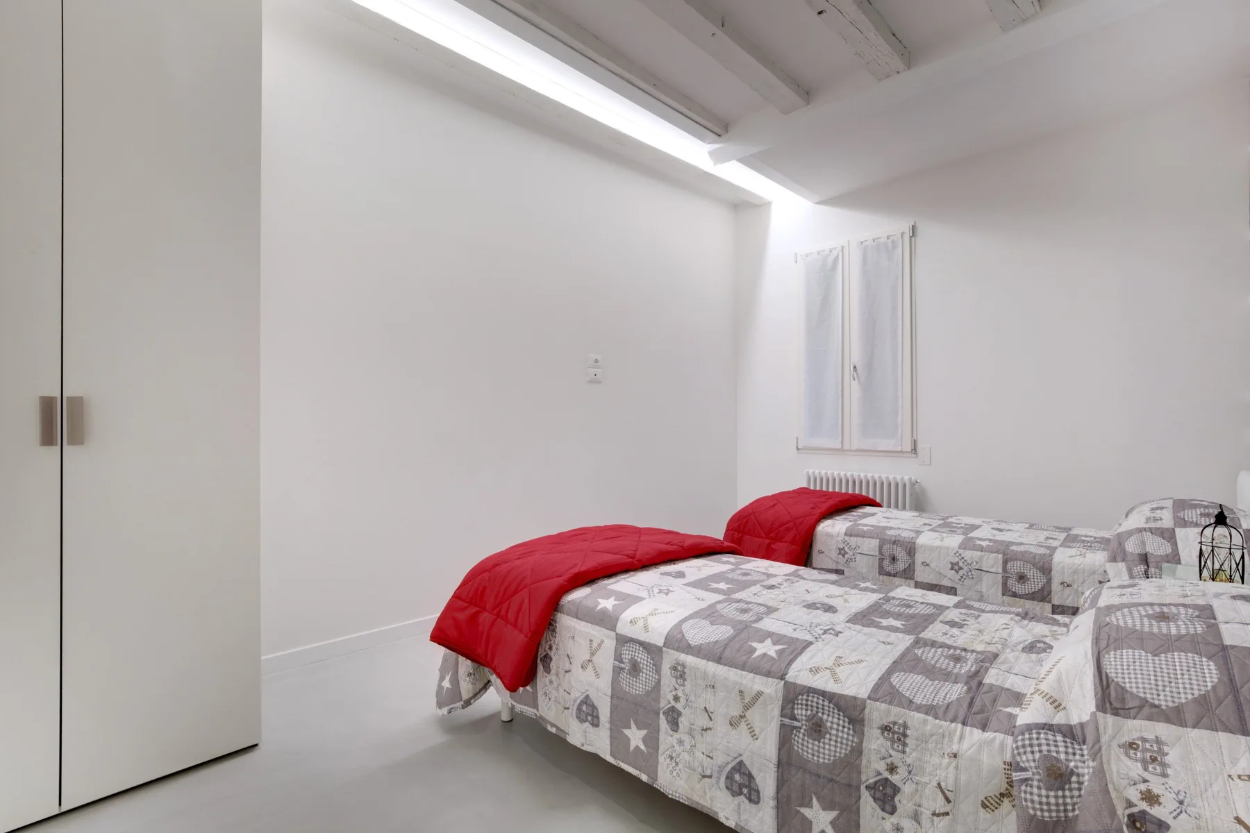 A bright bedroom has two beds with patterned bedding and red blankets.