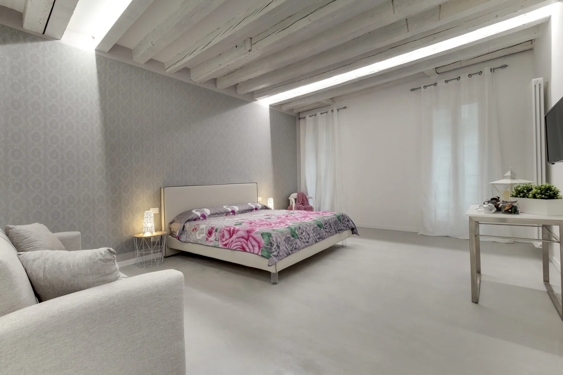 Un dormitorio con una cama cubierta con una colcha floral y un sillón de color claro.