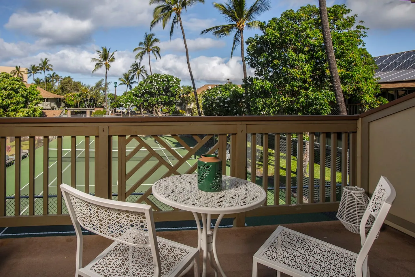 A patio seating area overlooks a tennis court, with palm trees beyond the railing.