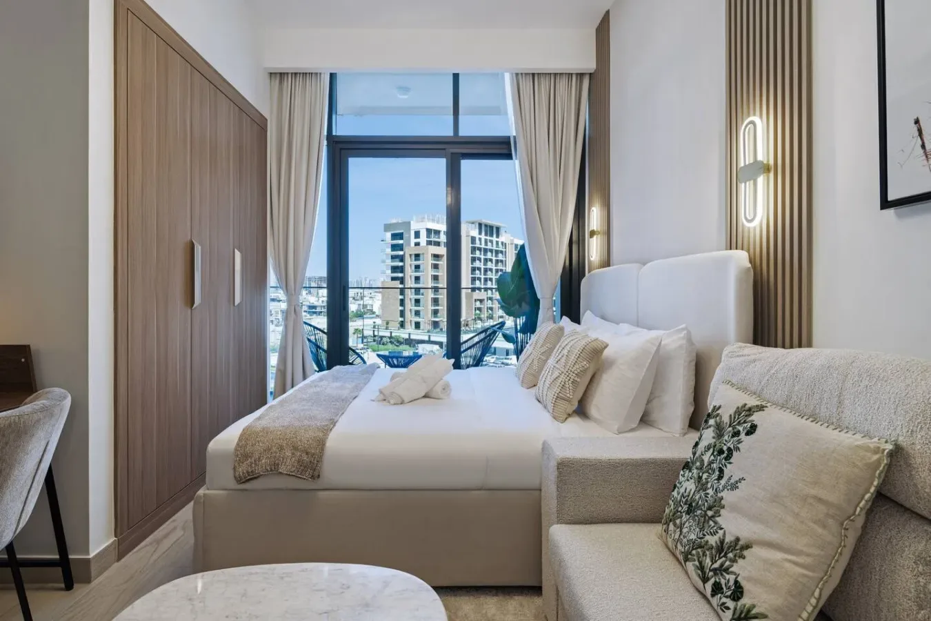 Un dormitorio con una cama bien hecha y grandes ventanas con vistas a edificios cercanos.