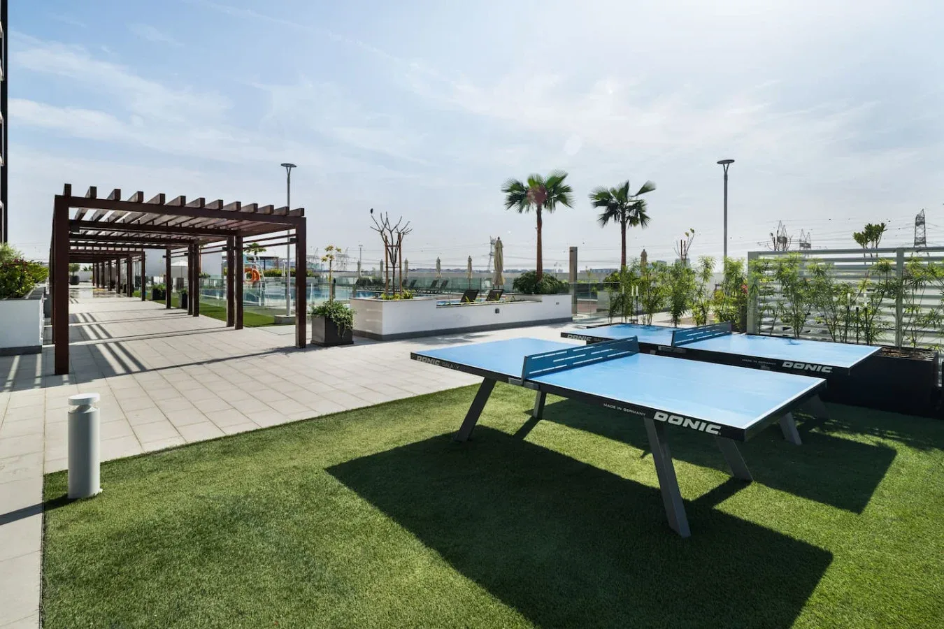 Una zona recreativa al aire libre tiene mesas de ping-pong junto a una pérgola y un jardín.