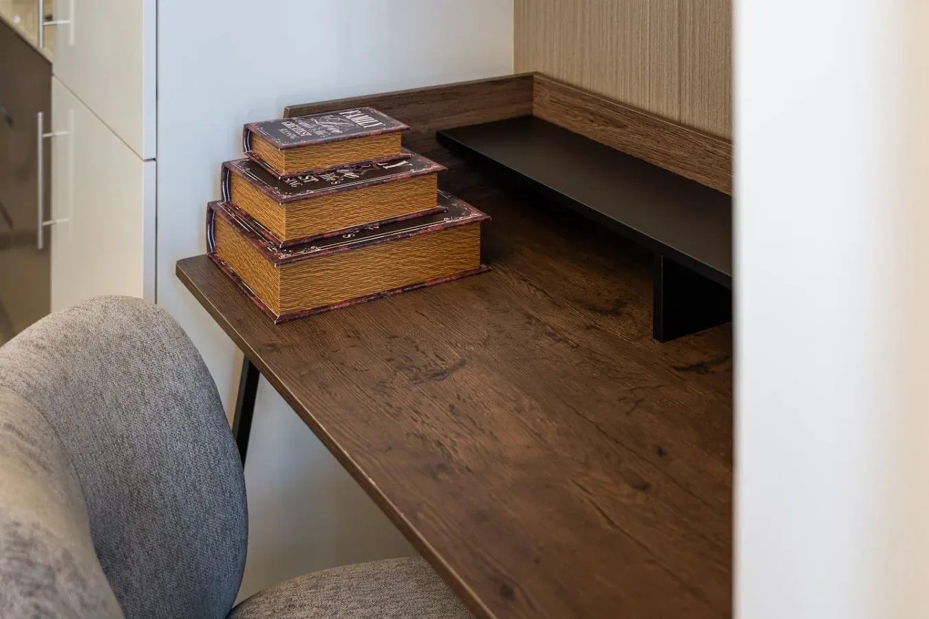Un escritorio de madera con libros apilados está junto a una silla tapizada gris.