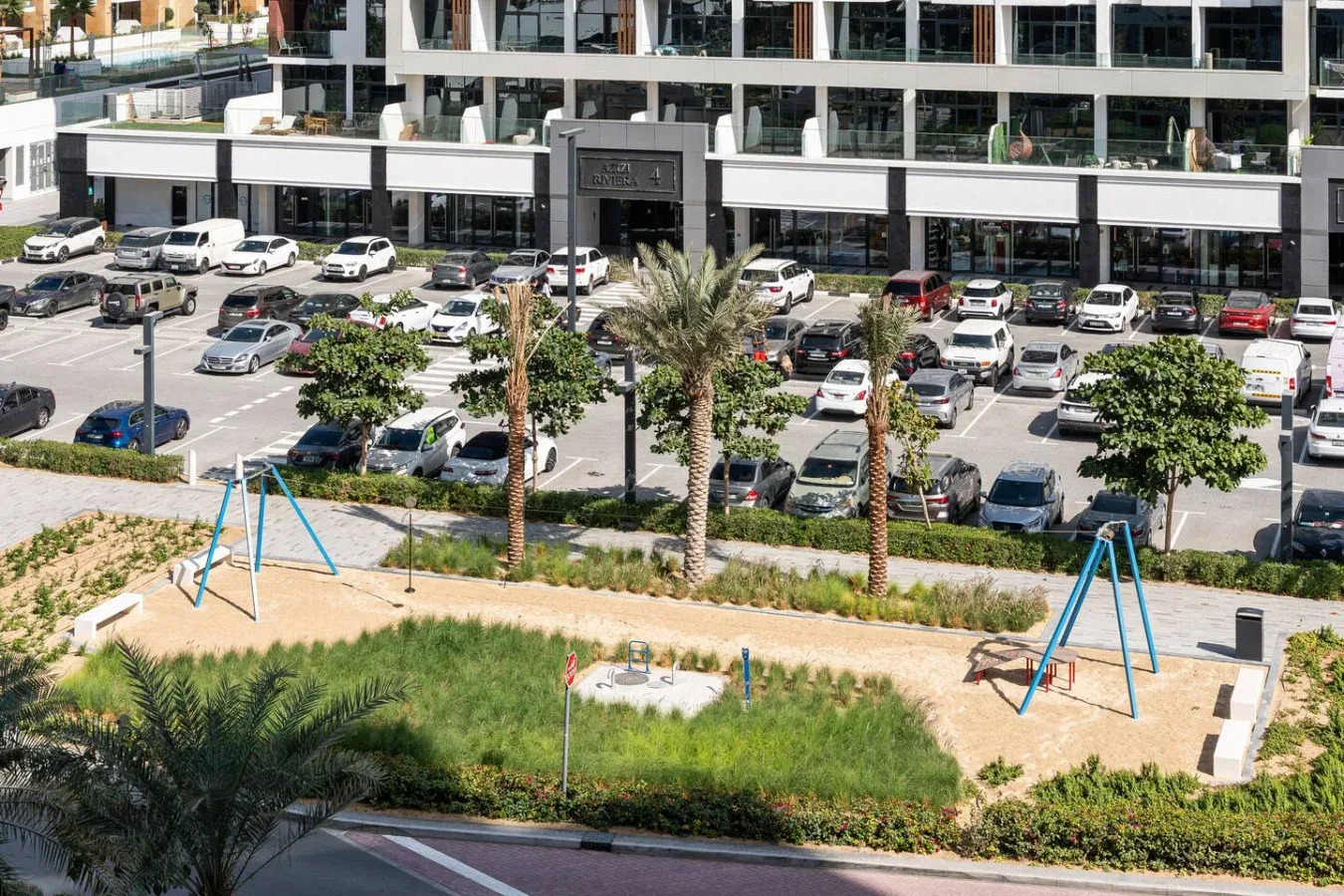 Un pequeño parque infantil con columpios está junto a un estacionamiento y un edificio moderno.