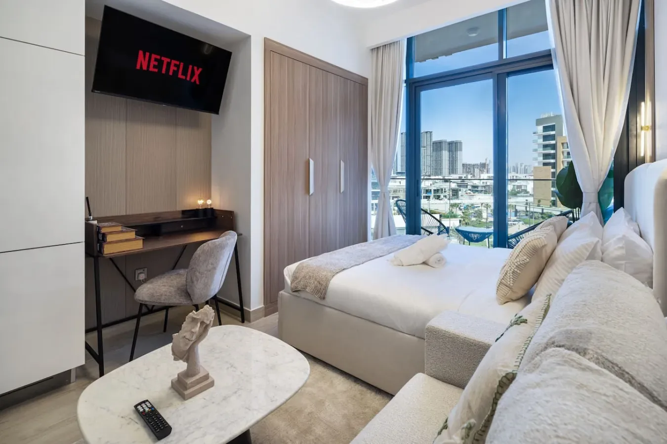 Dormitorio luminoso con cama, escritorio y grandes ventanas con edificios a la vista.