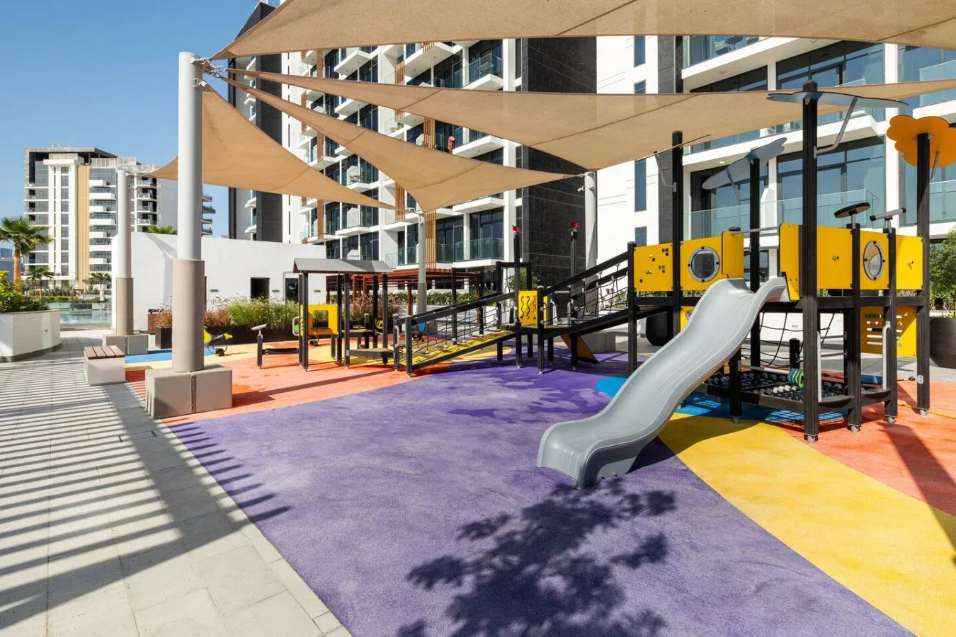 Un parque infantil al aire libre con sombra, un tobogán y suelo de colores.