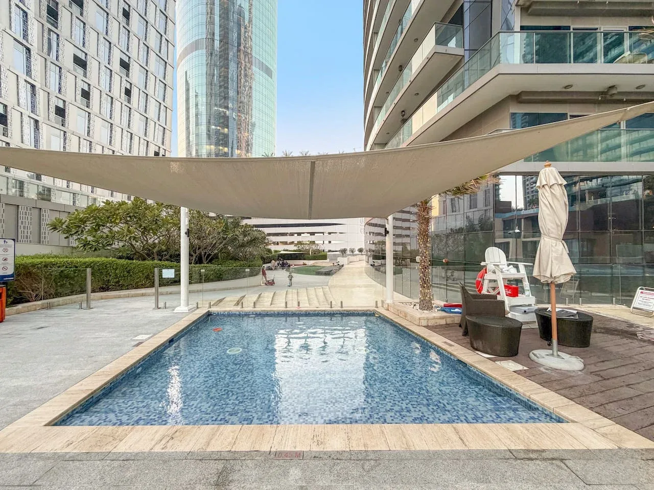 Una piscina al aire libre está bajo una gran cubierta de sombra entre edificios modernos.