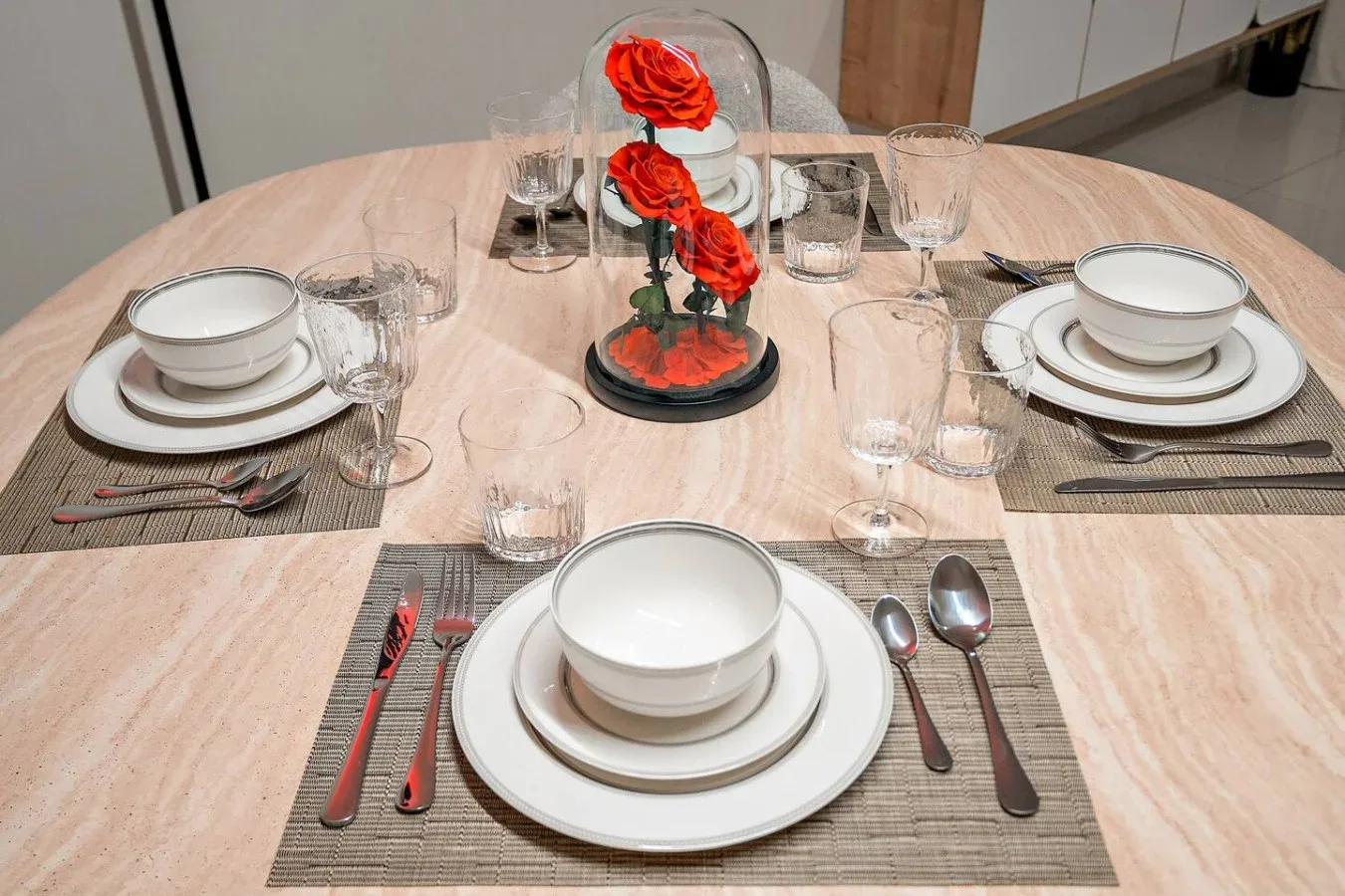Una mesa de comedor está puesta con platos, vasos y un centro floral rojo.