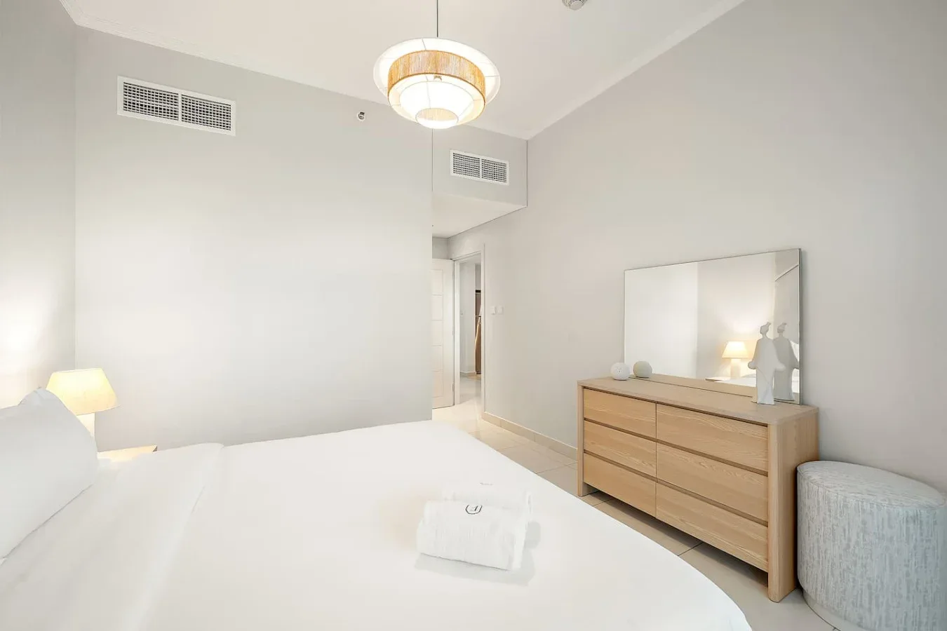 Un dormitorio luminoso con una cama bien hecha y una cómoda de madera bajo un espejo.