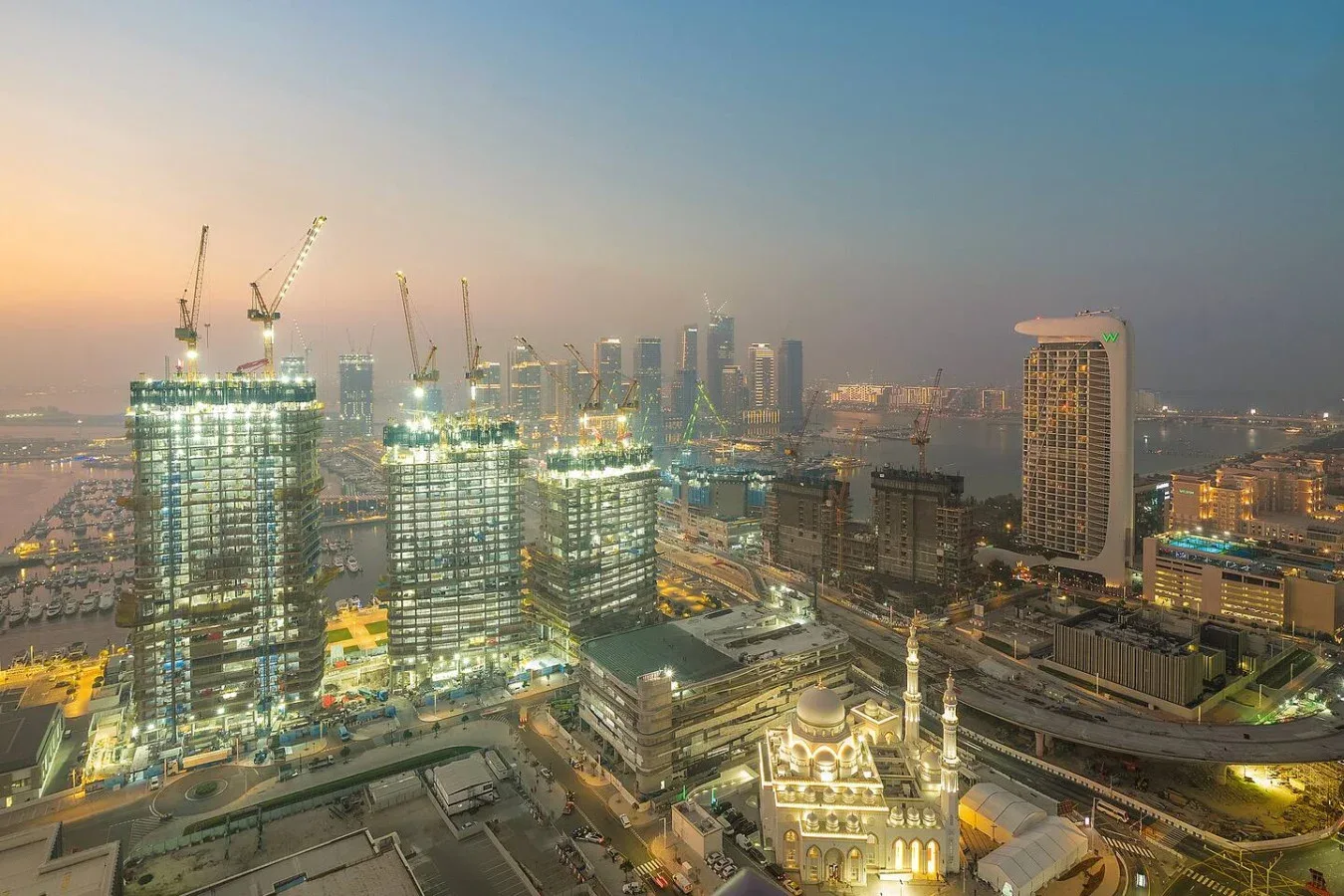 Un horizonte urbano al atardecer con edificios altos iluminados y grúas de construcción.