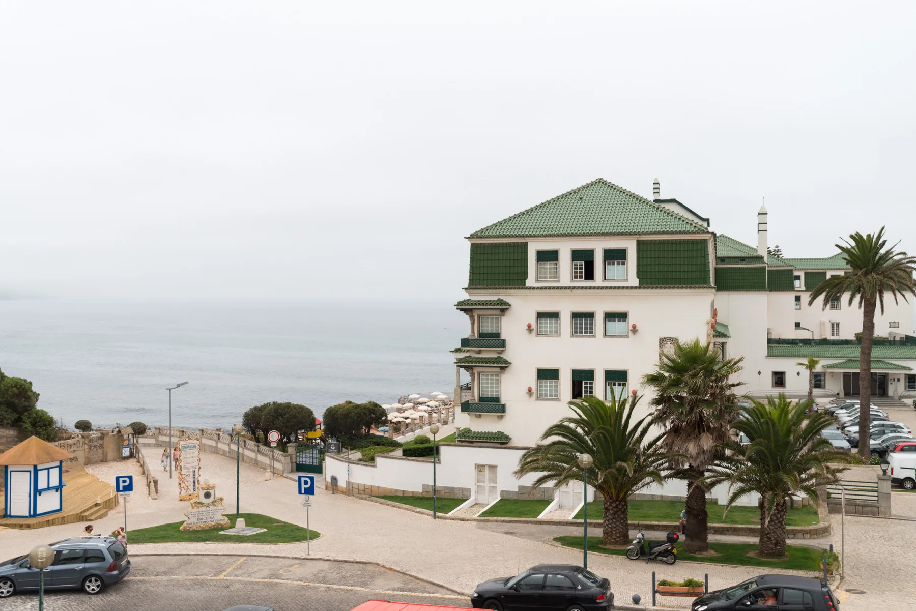 A coastal street overlooks the ocean beside a green-trimmed building and palm trees.