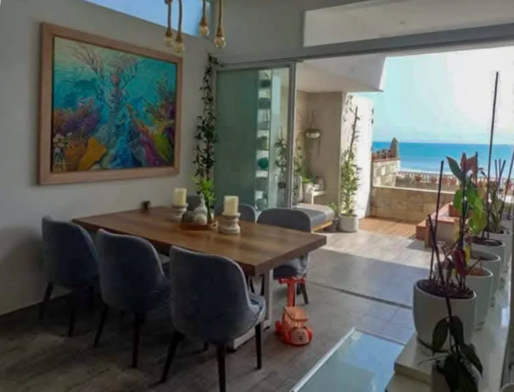 A dining area with a table and seating beside an open terrace overlooking the sea.