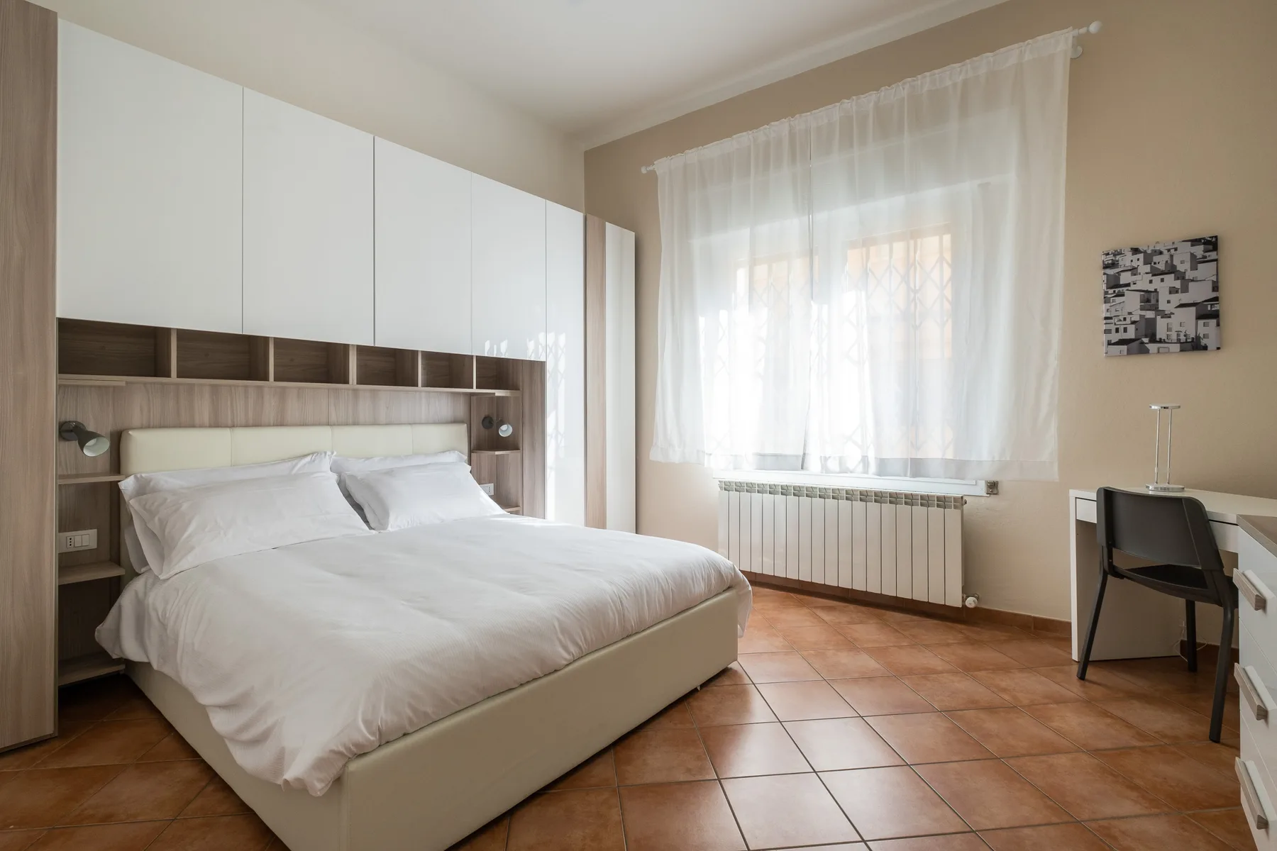 Un dormitorio luminoso con una cama bien arreglada y un escritorio junto a la ventana.