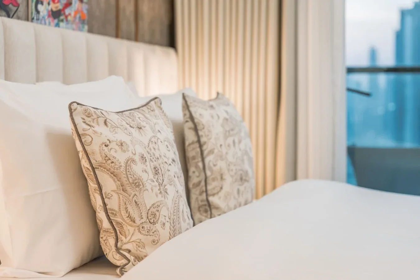 Una cama tendida con cojines estampados está junto a una ventana con cortinas.