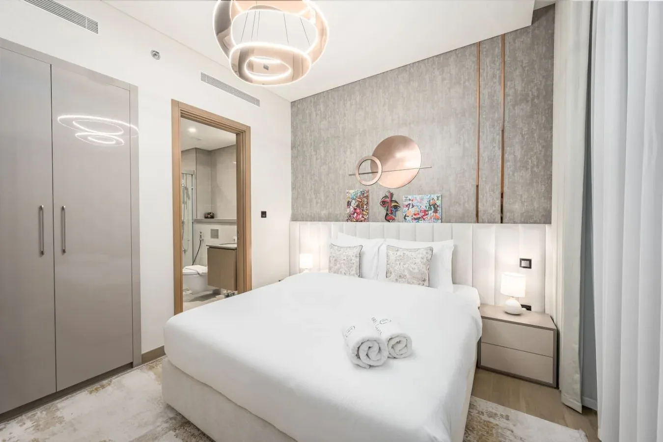 Un dormitorio luminoso con una cama blanca, mesitas de noche y una pared decorativa gris.