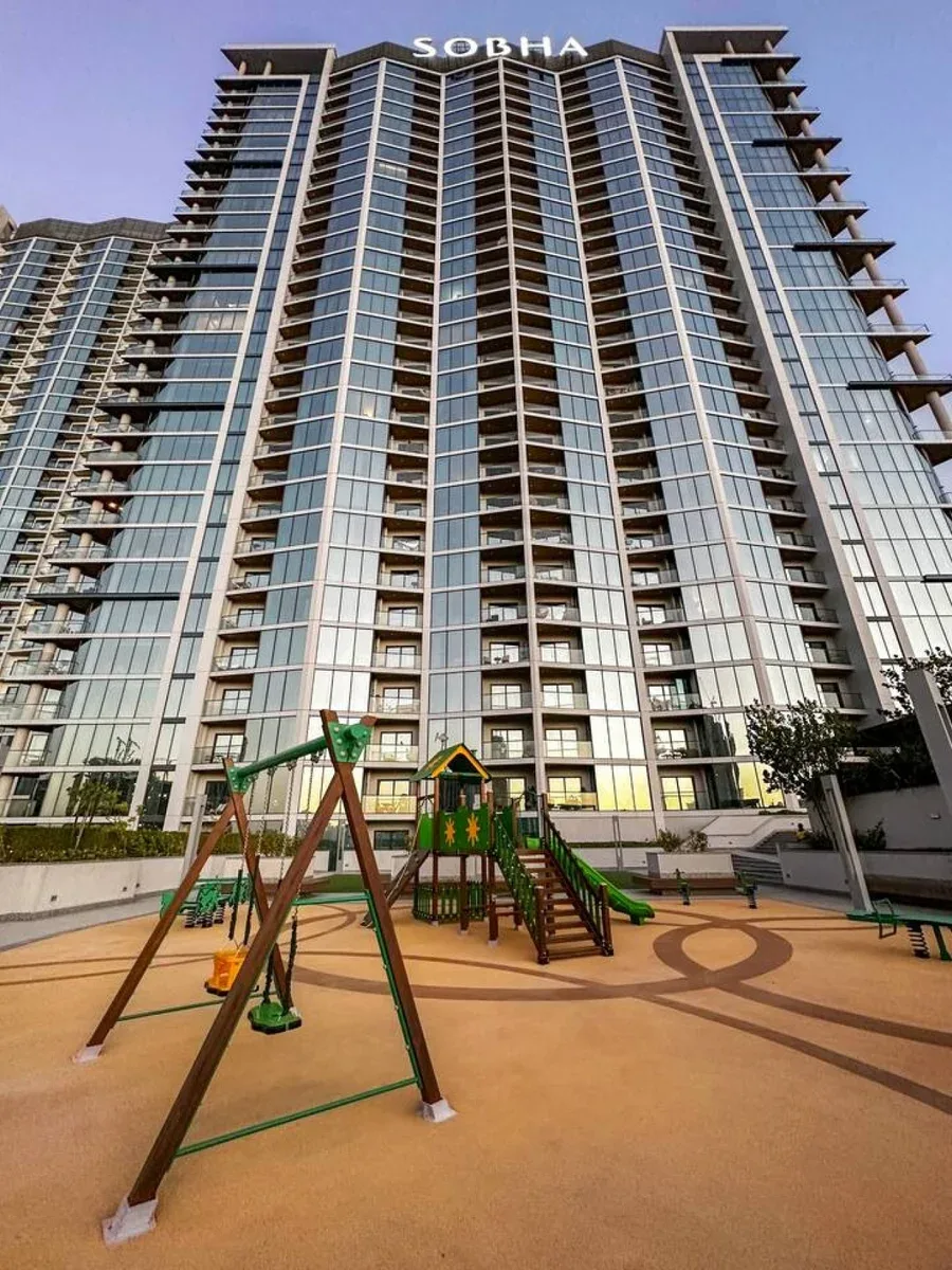 Una torre residencial alta se alza detrás de un parque infantil al aire libre con columpios y toboganes.