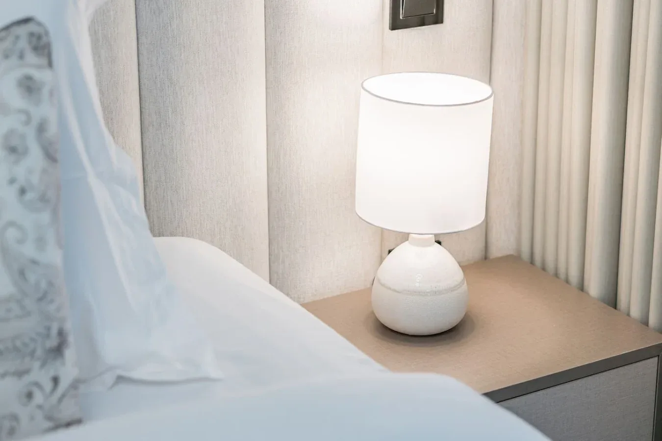 Una mesa de noche tiene una lámpara blanca junto a una cama.