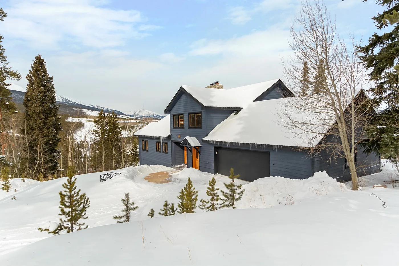 A dark-colored house sits in a snowy landscape beneath a partly cloudy sky.