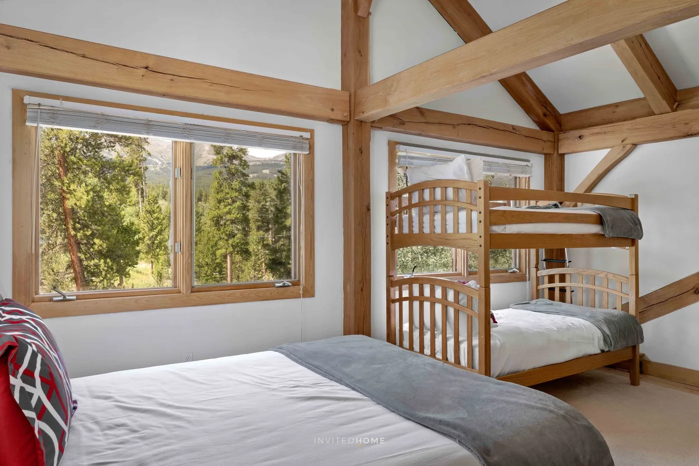 A bedroom with beds beneath exposed wooden beams and windows overlooking trees.