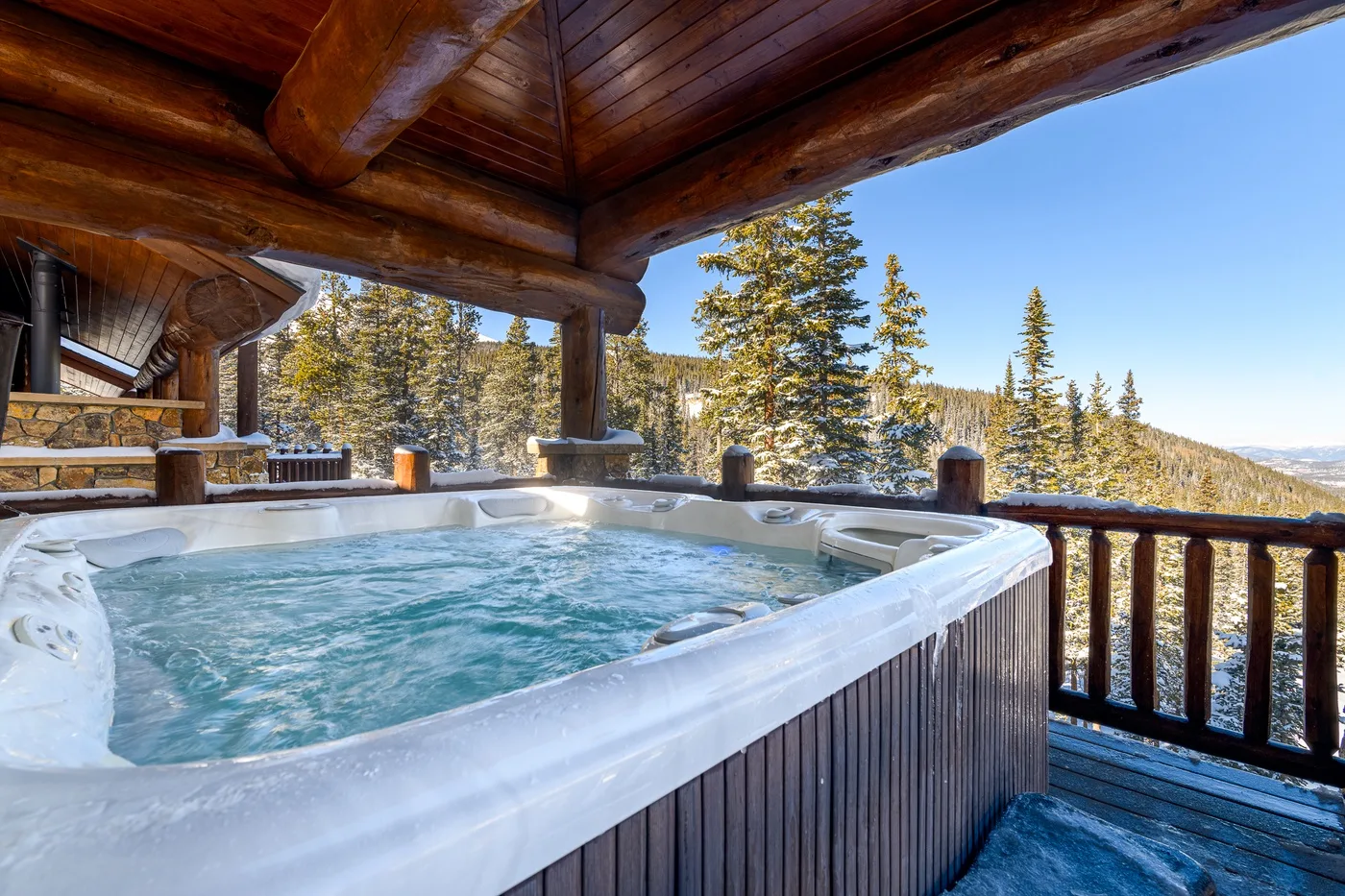A covered outdoor hot tub sits beside a wooden railing with pine trees beyond.