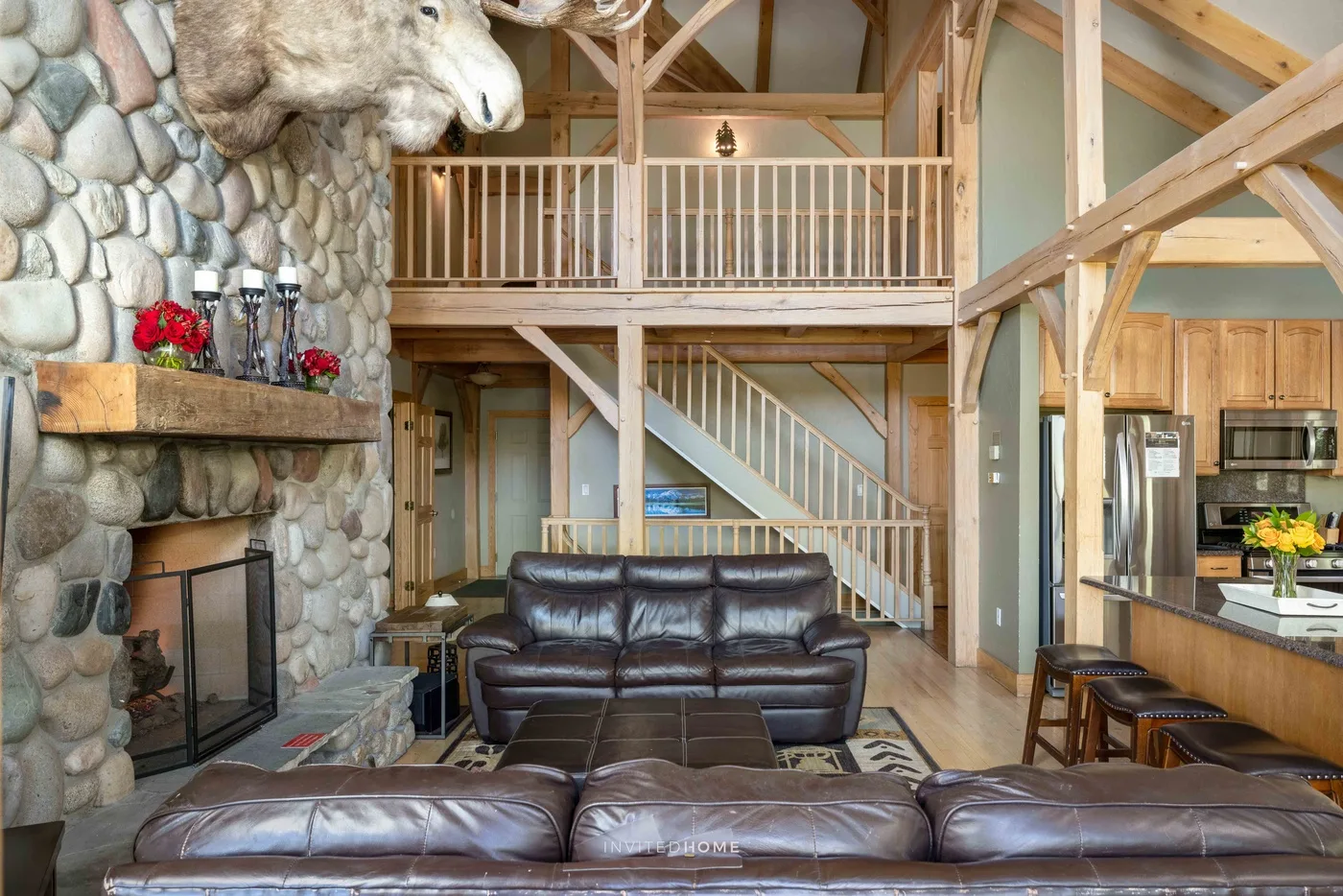 A spacious living room with leather seating, a stone fireplace, and a wooden loft railing.