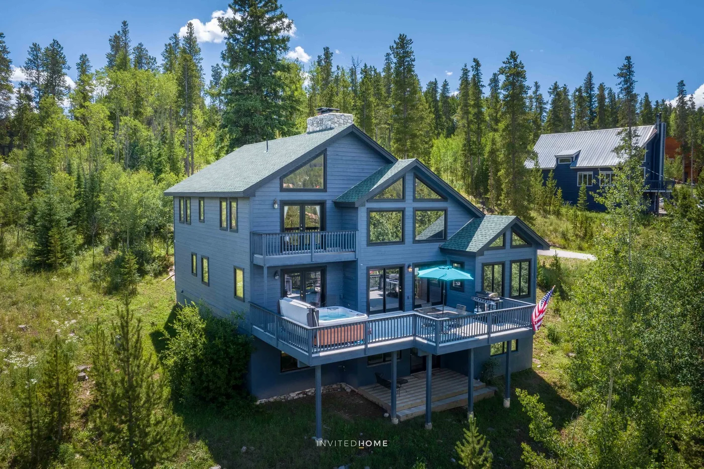 A large house with a wraparound deck stands among pine trees.