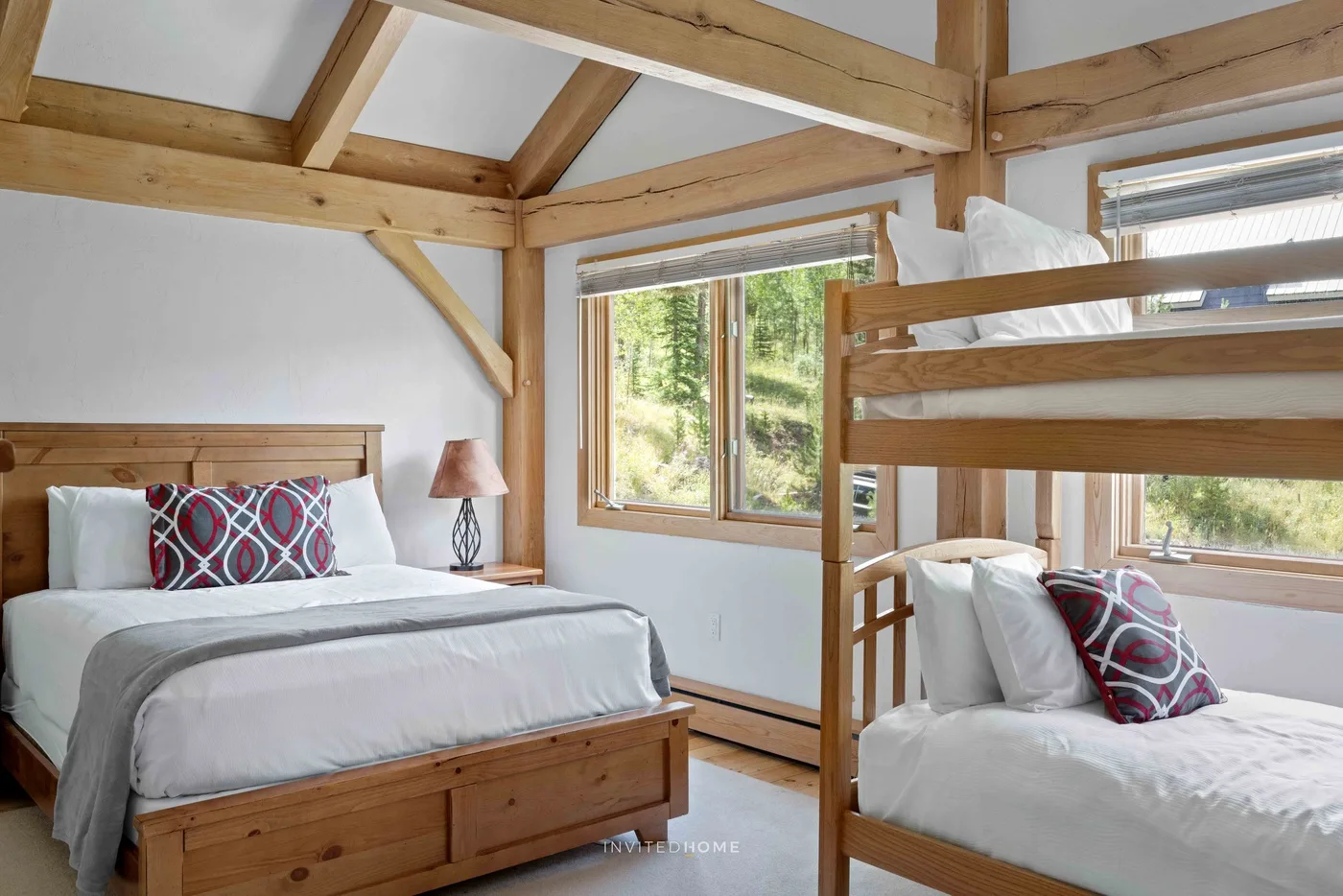 A bedroom with two beds, exposed wooden beams, and windows overlooking trees.
