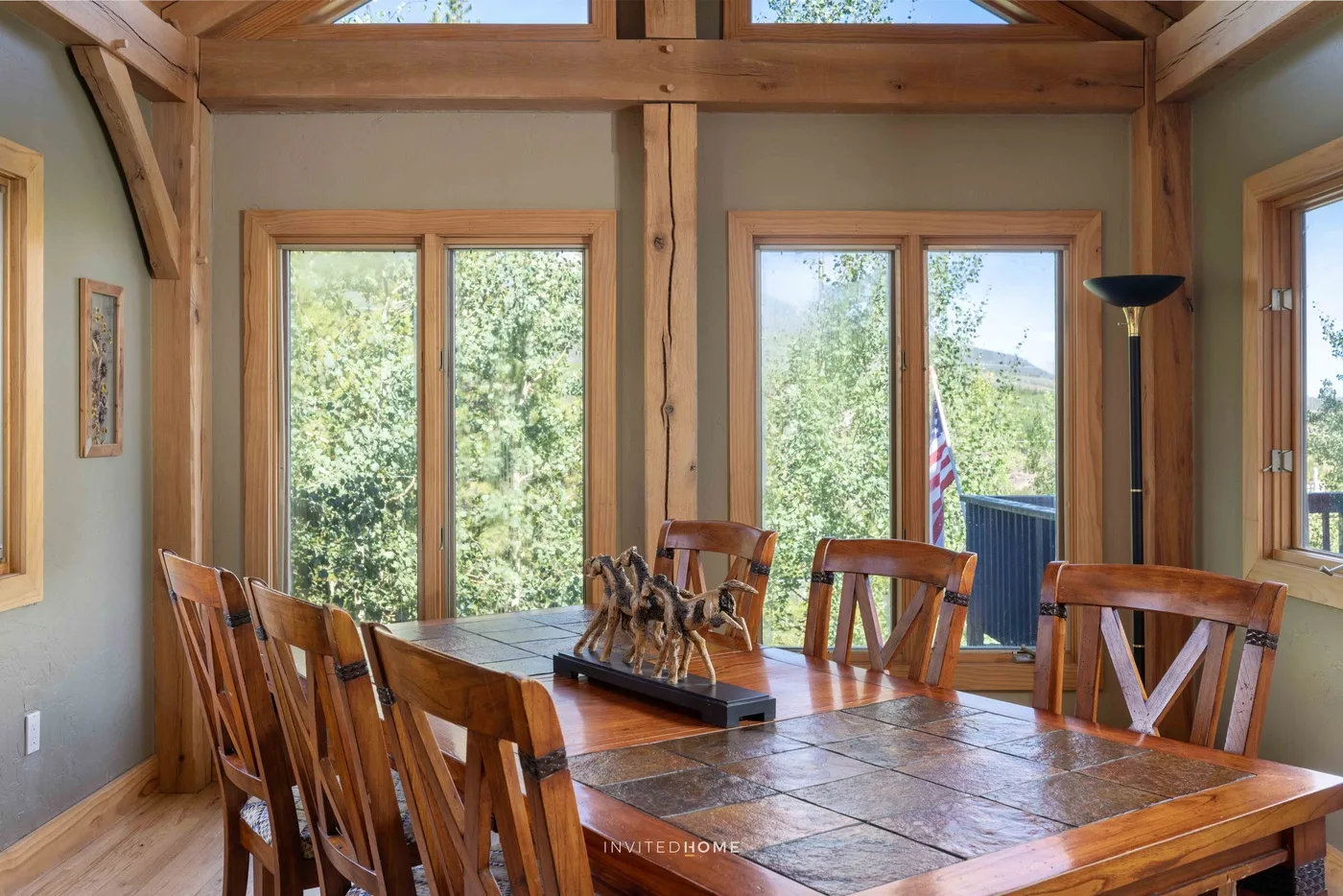 A wooden dining table and seating sit beside large windows overlooking trees.