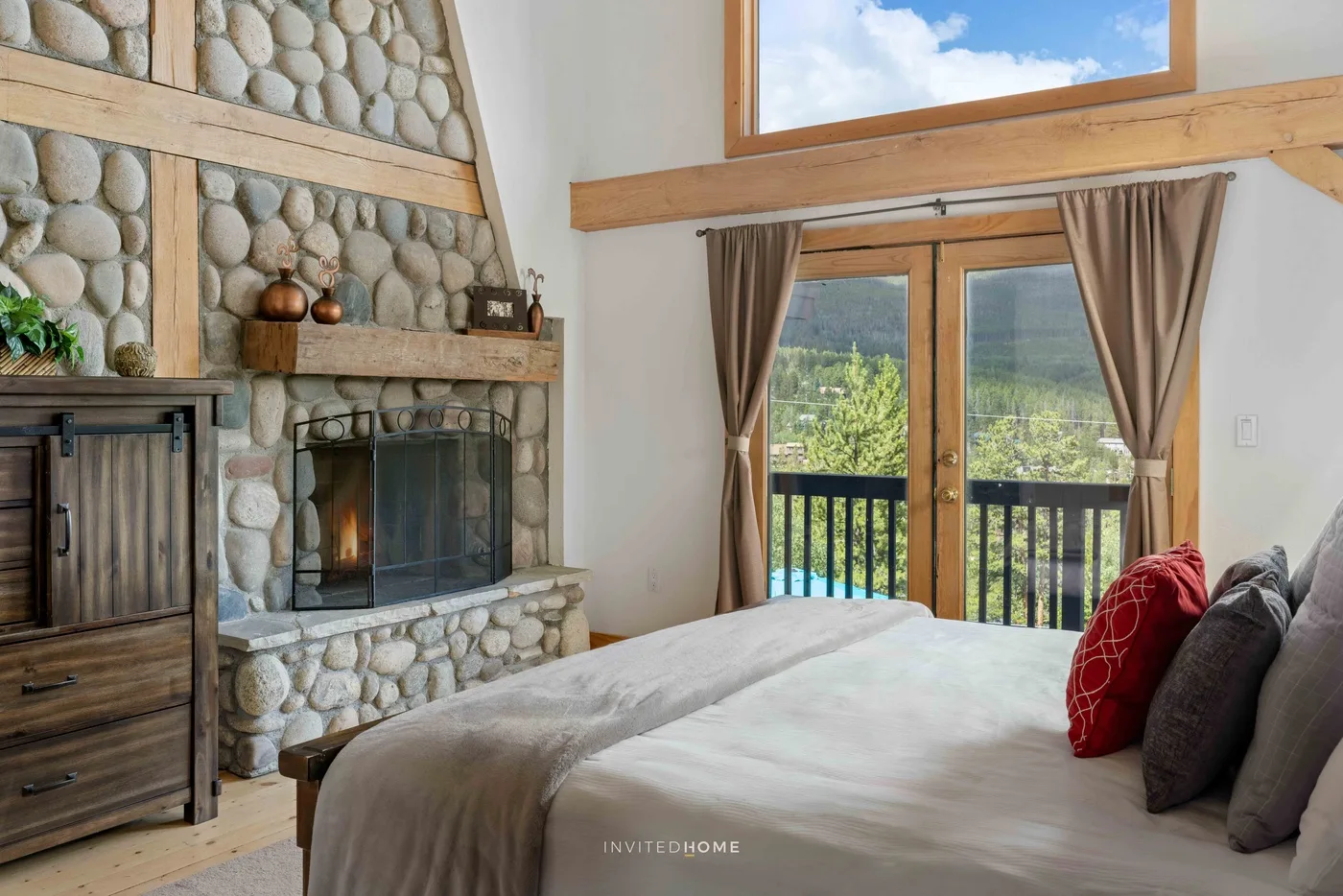 A bedroom with a bed beside a stone fireplace and glass doors opening to a balcony.
