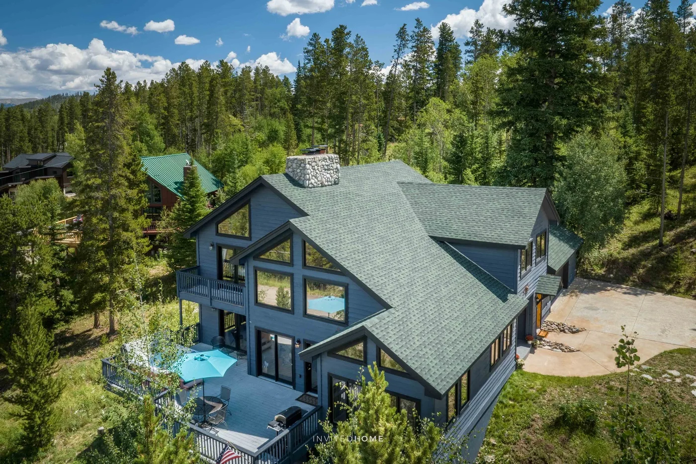 A modern cabin sits among tall trees, with a deck and turquoise umbrella.