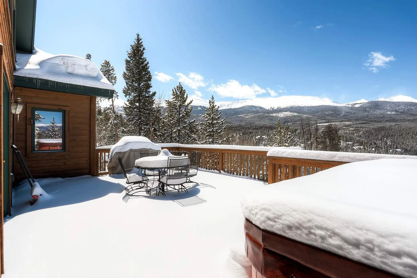 A snow-covered outdoor deck overlooks a mountain landscape, with seating beside the wooden building.