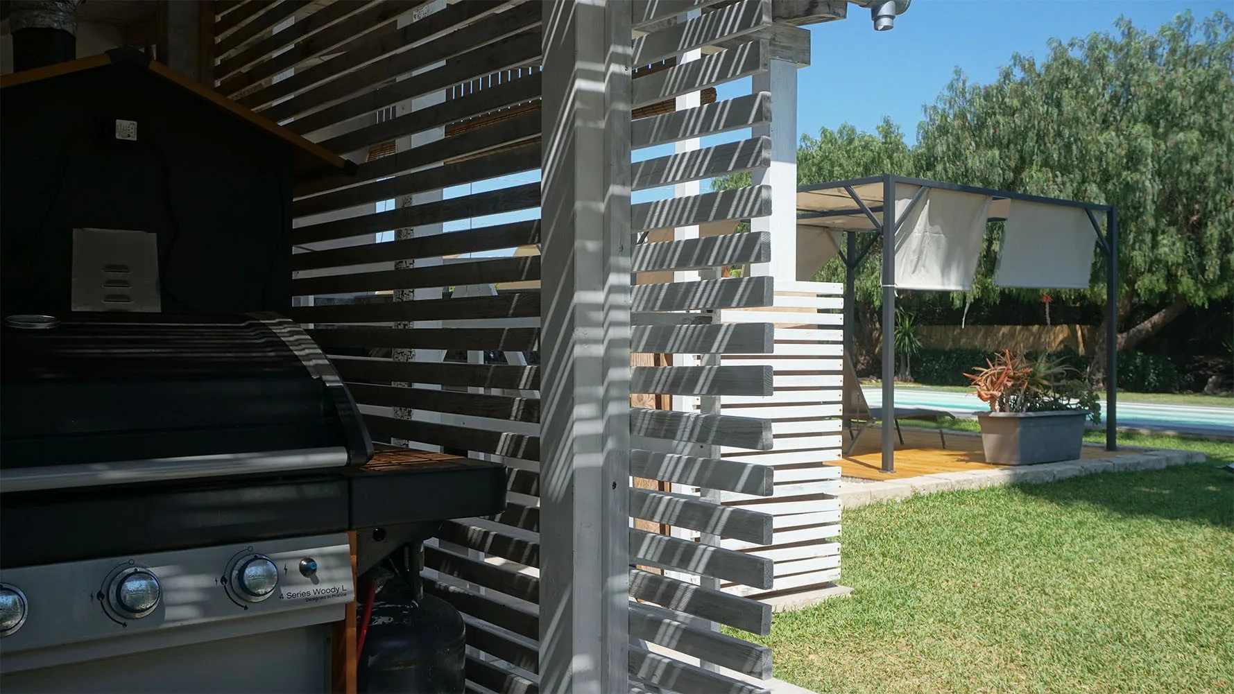 Una zona de parrilla al aire libre y techada da a un jardín con césped y piscina.