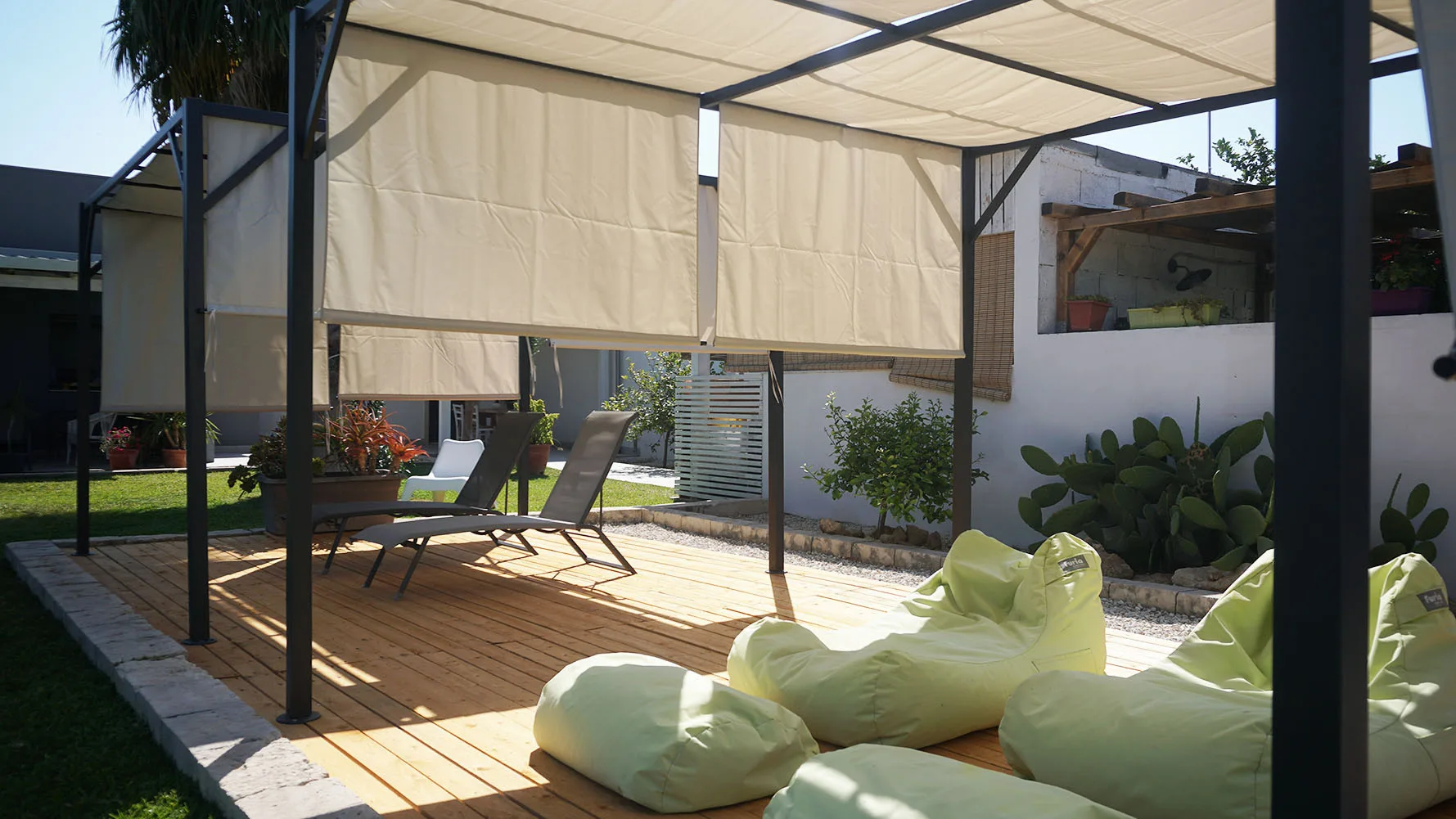 Un patio exterior cubierto tiene asientos bajos, toldos y plantas en macetas.
