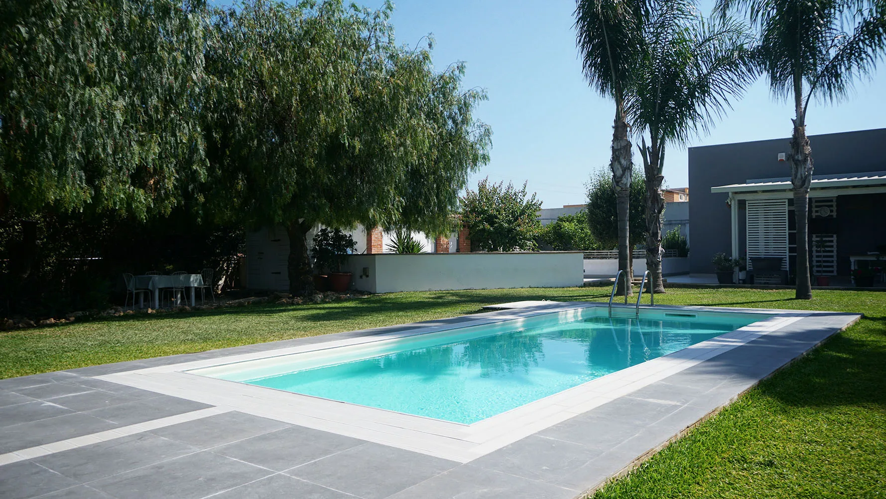 Piscina al aire libre rodeada de pavimento, césped y árboles.