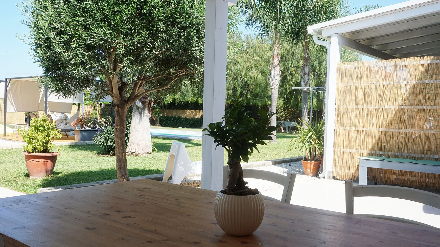 Un patio exterior a la sombra con una mesa de madera da a un jardín verde.