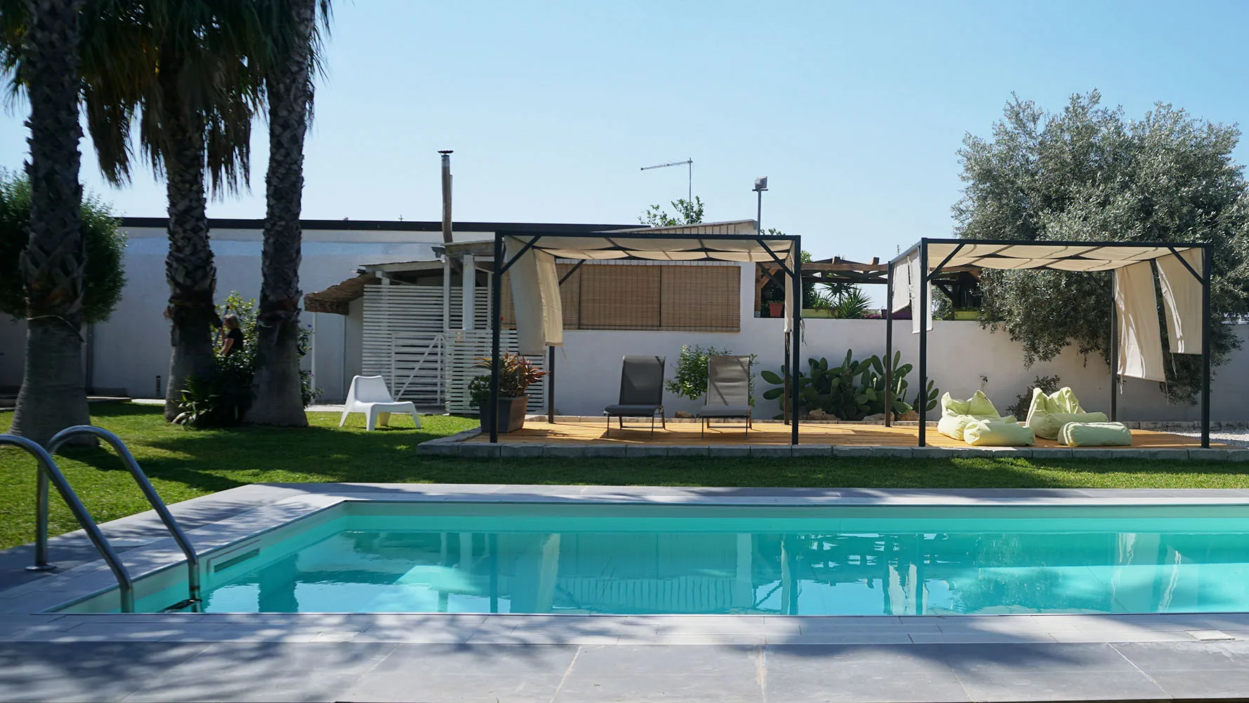 Una piscina al aire libre está frente a zonas de estar con sombra y un edificio bajo.
