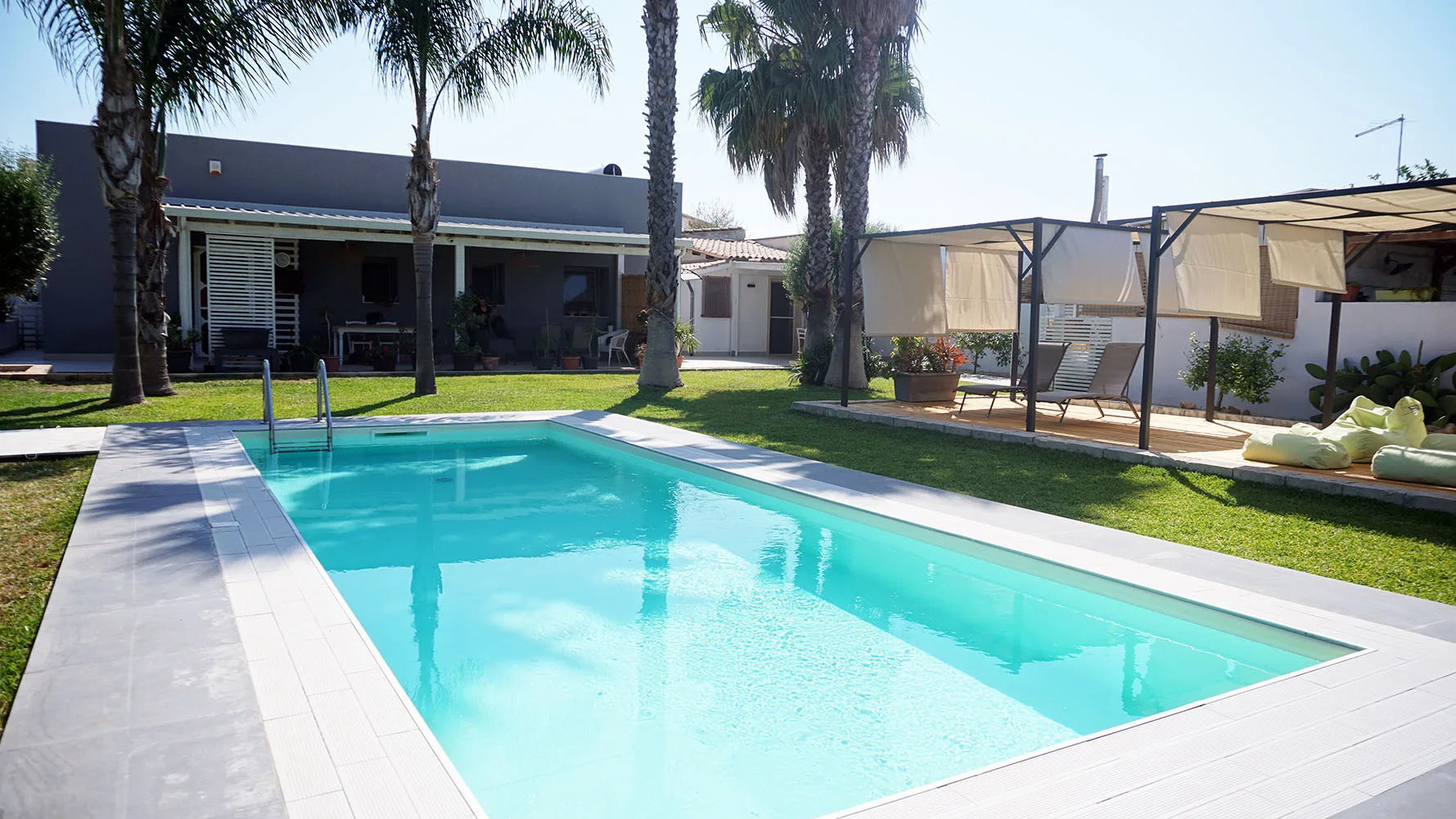 Una piscina al aire libre junto a un césped, palmeras y una zona de estar con sombra.