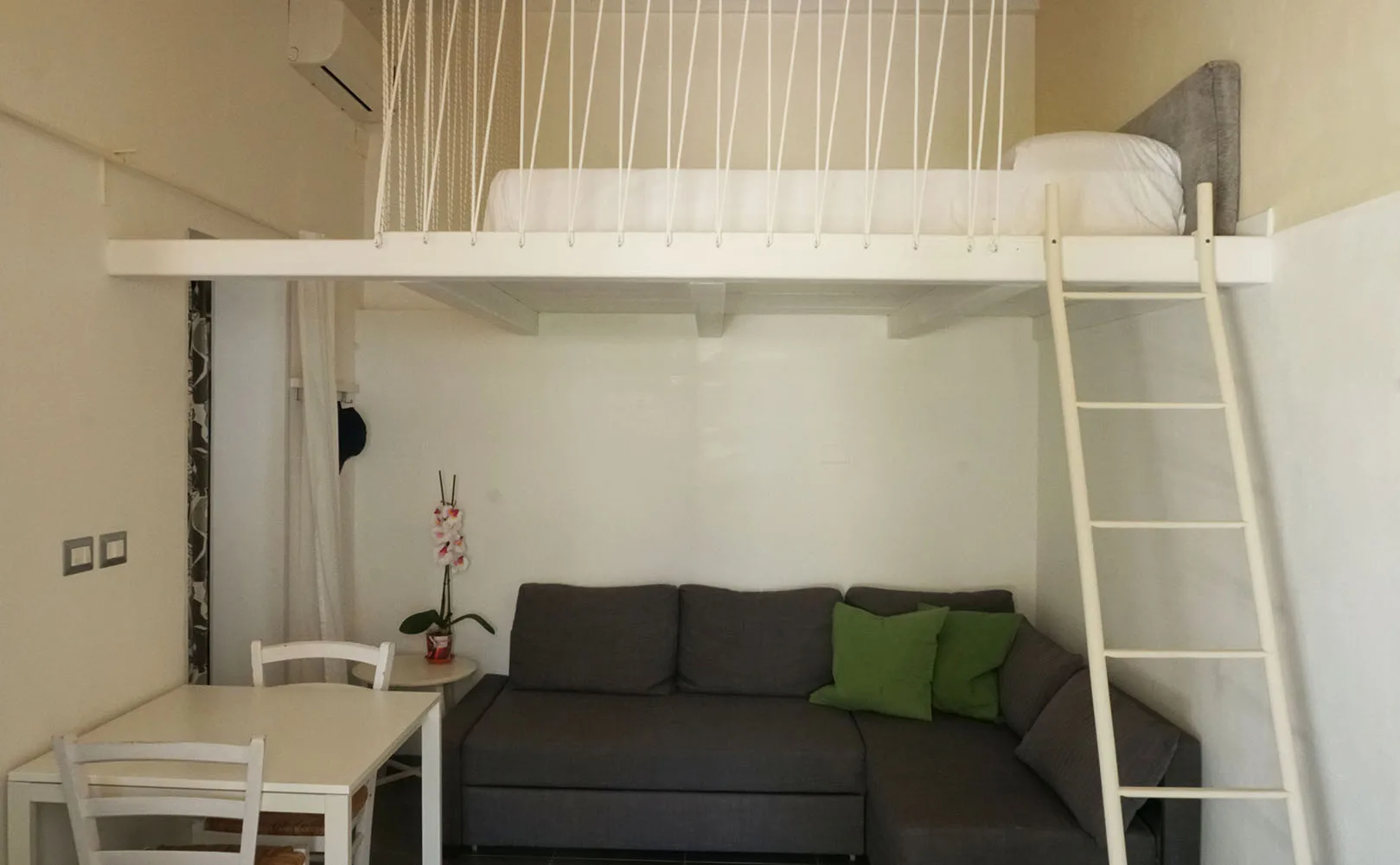 Una sala de estar con un sofá oscuro, cojines verdes y una mesa de comedor bajo una cama elevada.