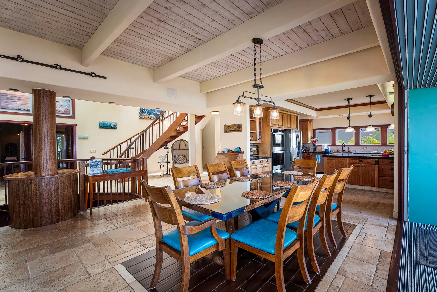 A dining area with a long wooden table and blue-cushioned seating beside an open kitchen.