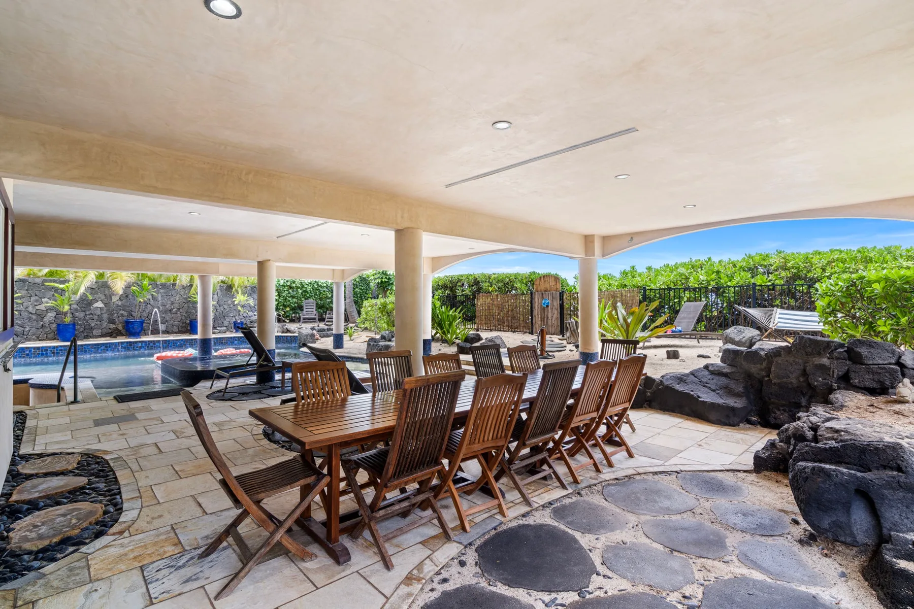A covered outdoor dining area with a long wooden table beside a swimming pool.