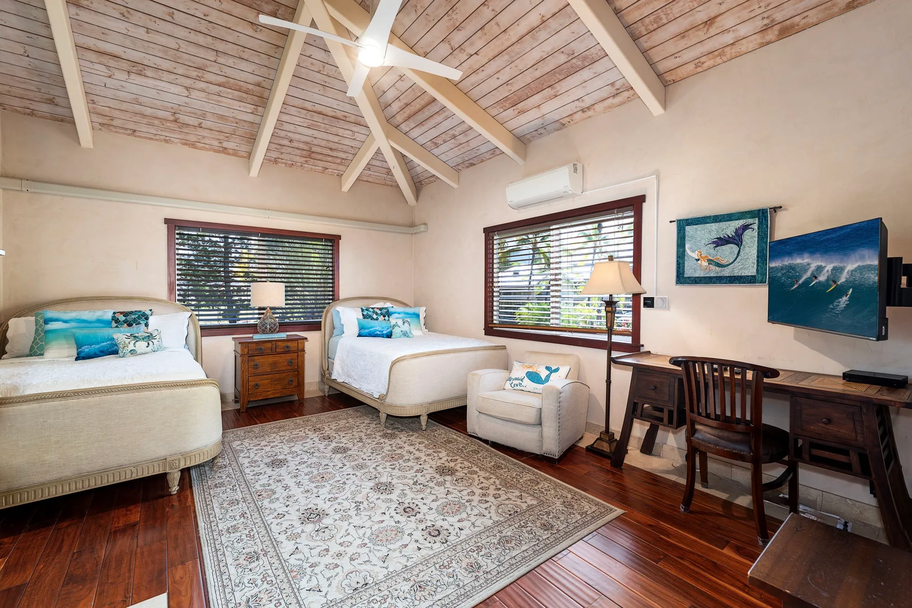 A bedroom seating area with two beds, an armchair, and a desk beneath a high wooden ceiling.
