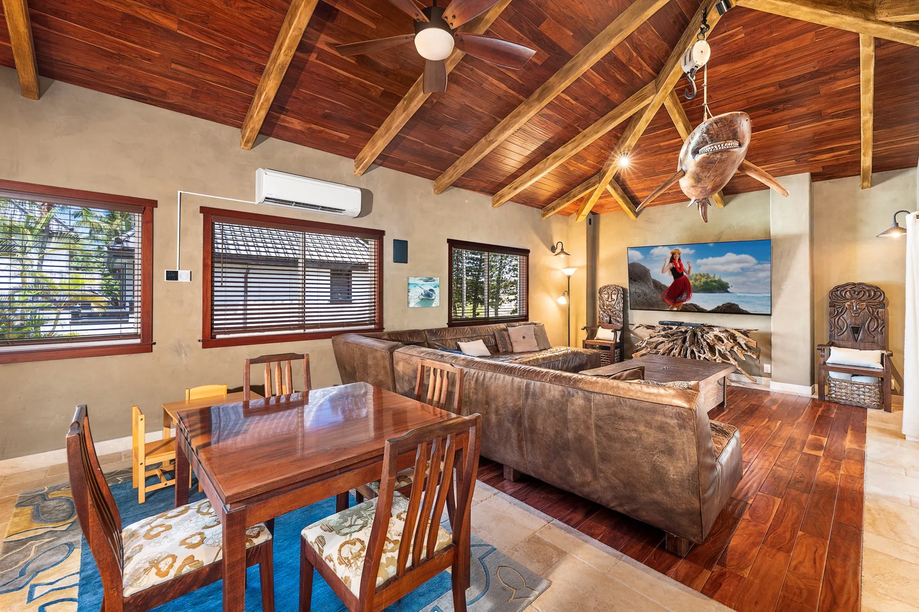 A living and dining room with wooden seating, large windows, and a peaked timber ceiling.