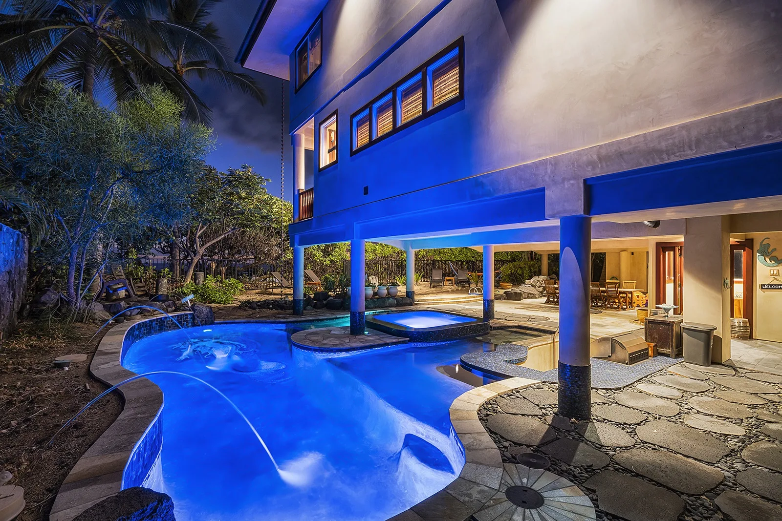 A brightly lit swimming pool sits beside a modern building and stone patio at night.
