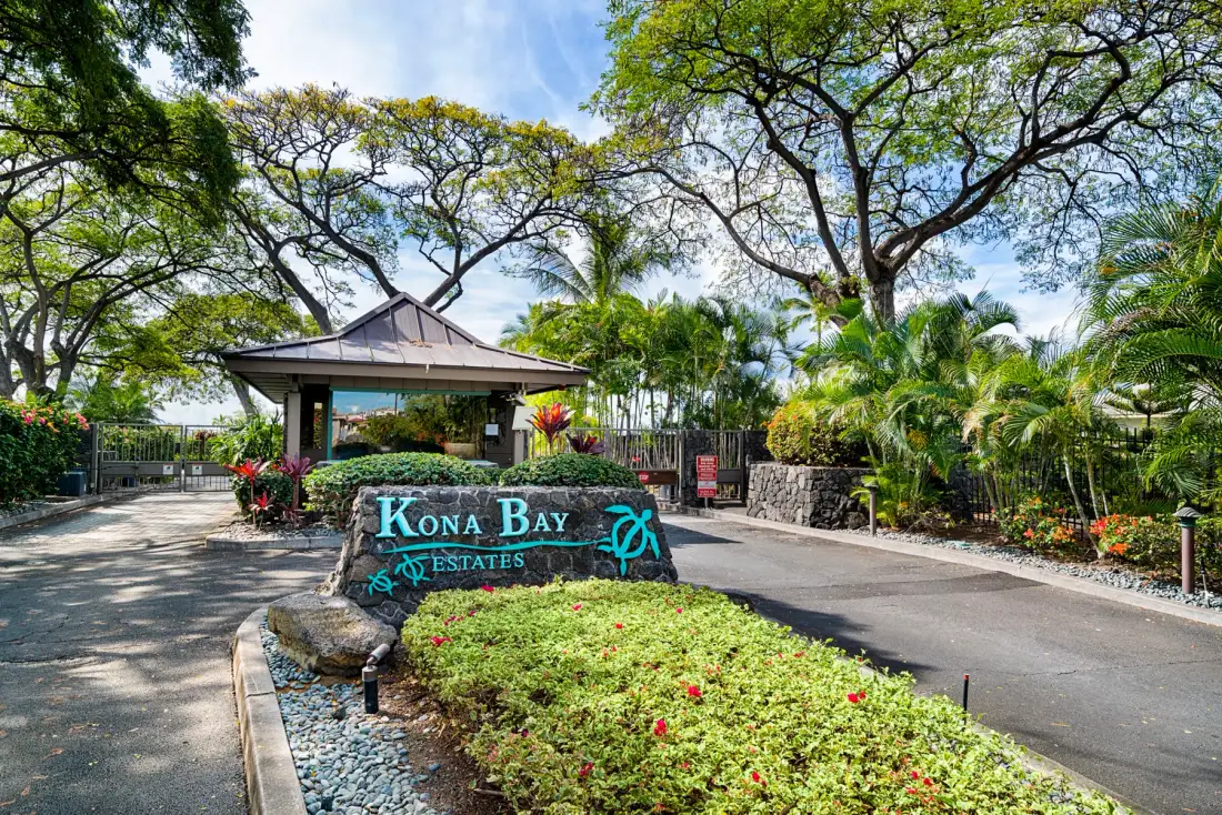 A landscaped entrance features a stone sign beneath large leafy trees.