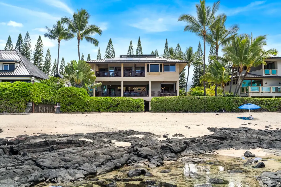 A beachfront house stands behind a sandy shore and dark rocks.