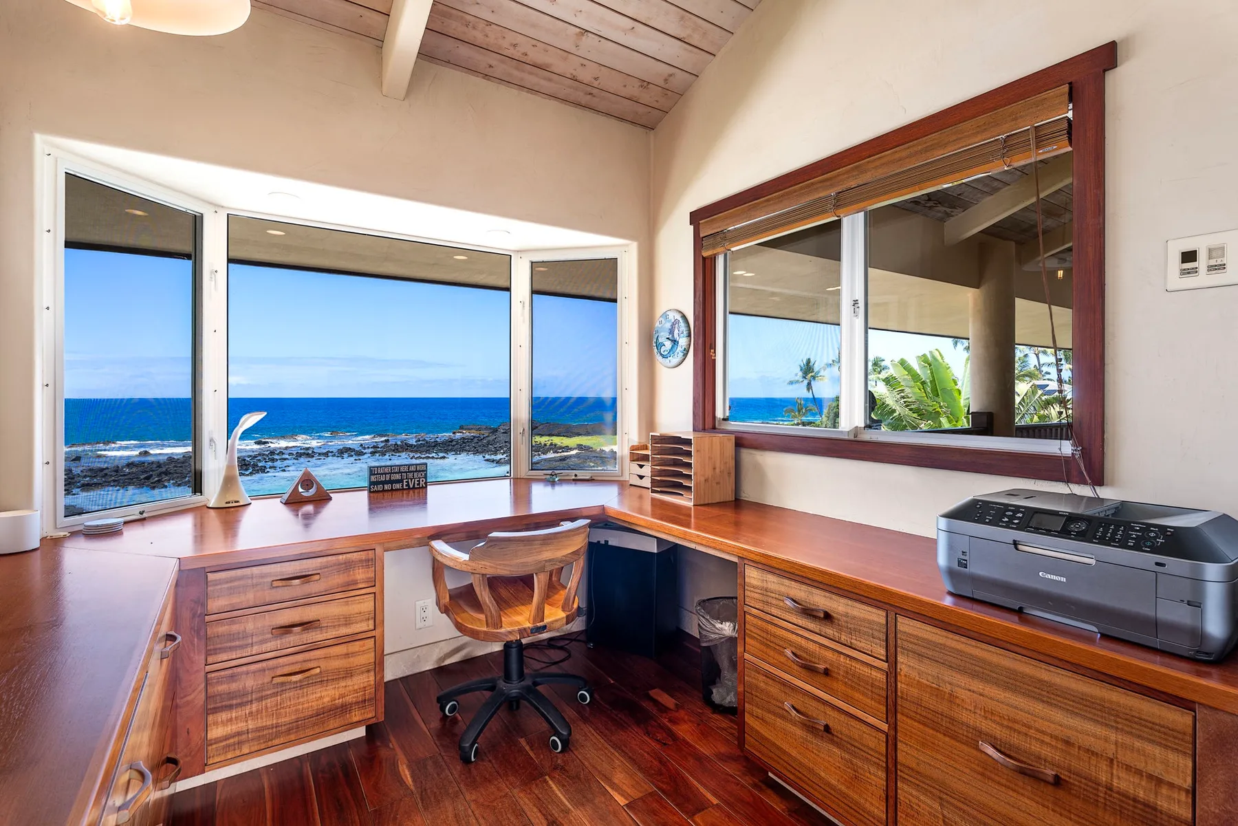 A wooden desk and office chair sit beside wide windows overlooking the ocean.